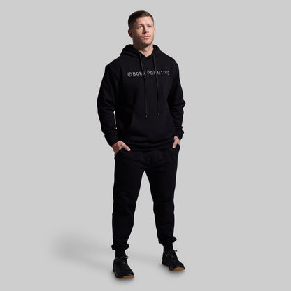 Man wearing a black hoodie and pants with a brand logo on a gray background