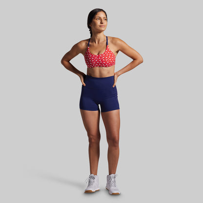 Woman wearing a red sports bra with white patterns and blue shorts on a gray background