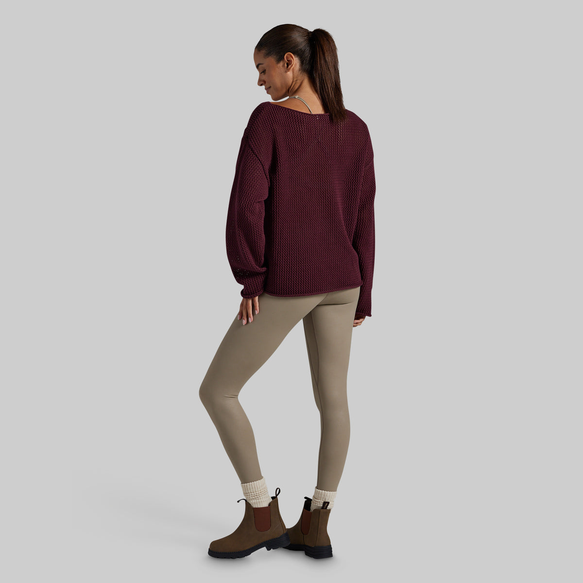 A woman shown from the back wearing a dark burgundy ribbed knit sweater, beige leggings, white socks, and brown Chelsea boots, standing against a solid light gray background.