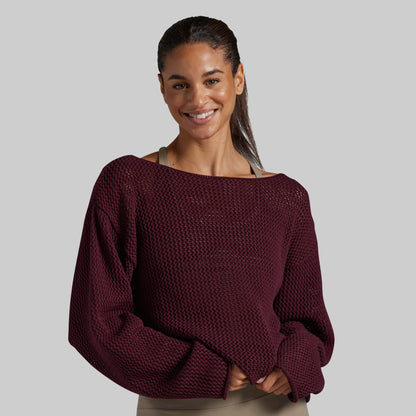 A woman wearing a textured, off-the-shoulder knit sweater in deep burgundy, photographed against a solid light gray studio background.