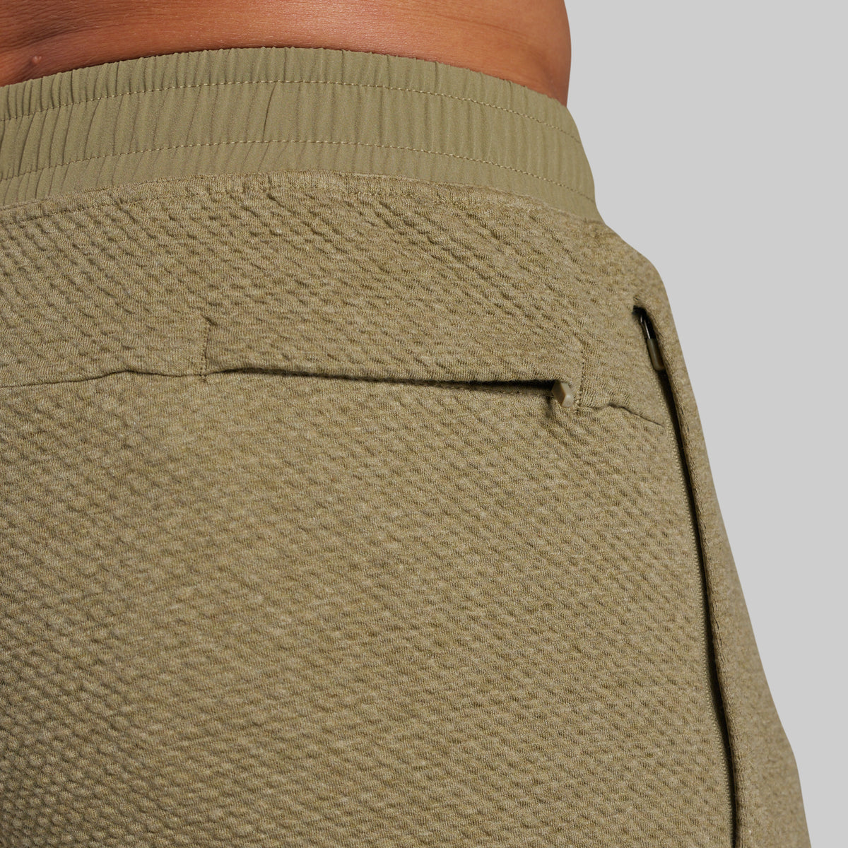 Close-up of the back waistband and pocket area of textured olive green athletic jorggers, showing a ribbed elastic waistband and a welt pocket on a light gray background.