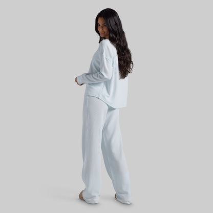A woman modeling a light blue lounge set consisting of a long-sleeve top and matching wide-leg pants, shown from a side-back angle against a solid gray studio background.