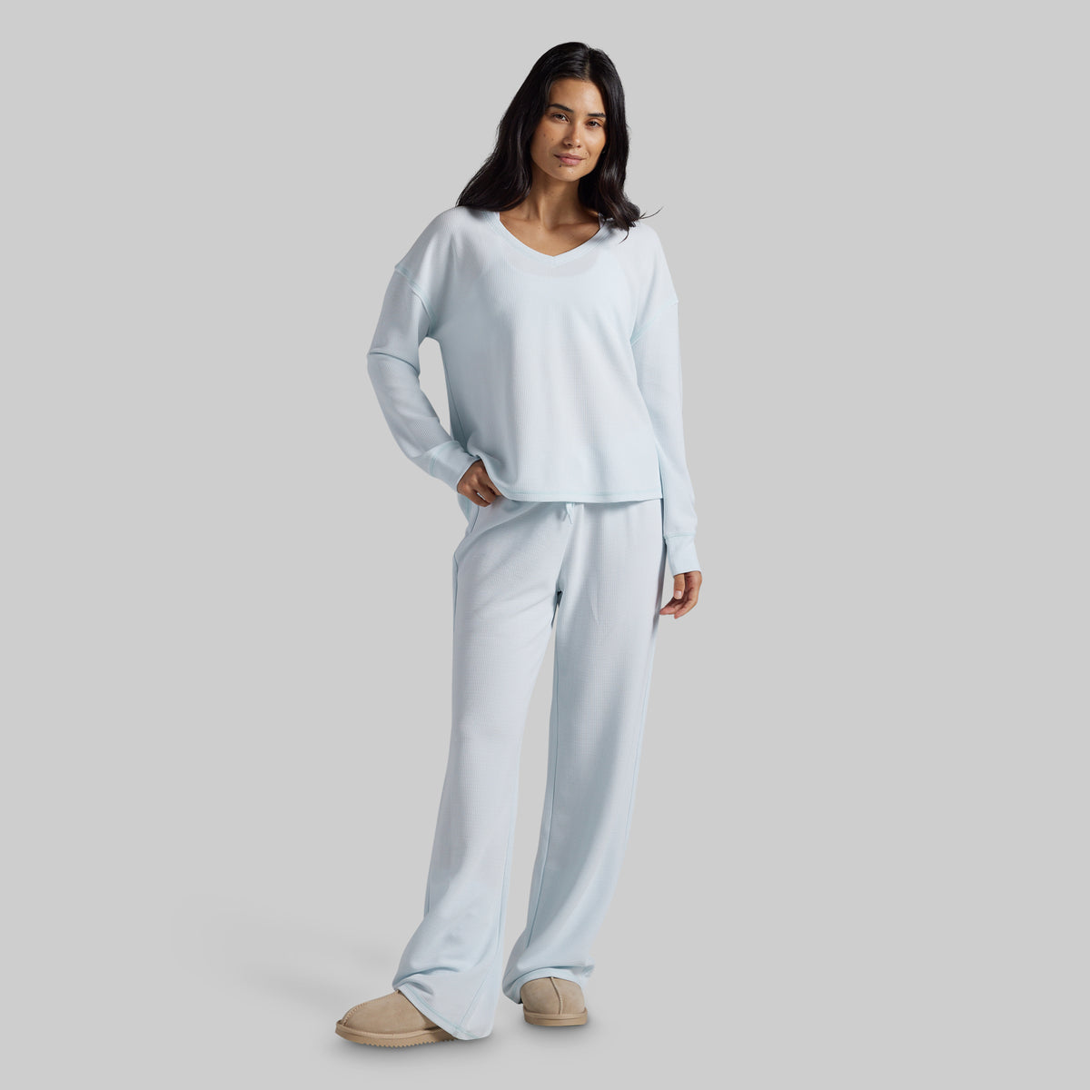 A woman modeling a light blue two-piece loungewear set consisting of a long-sleeve V-neck top and matching wide-leg pants, paired with beige slippers against a solid gray studio background.