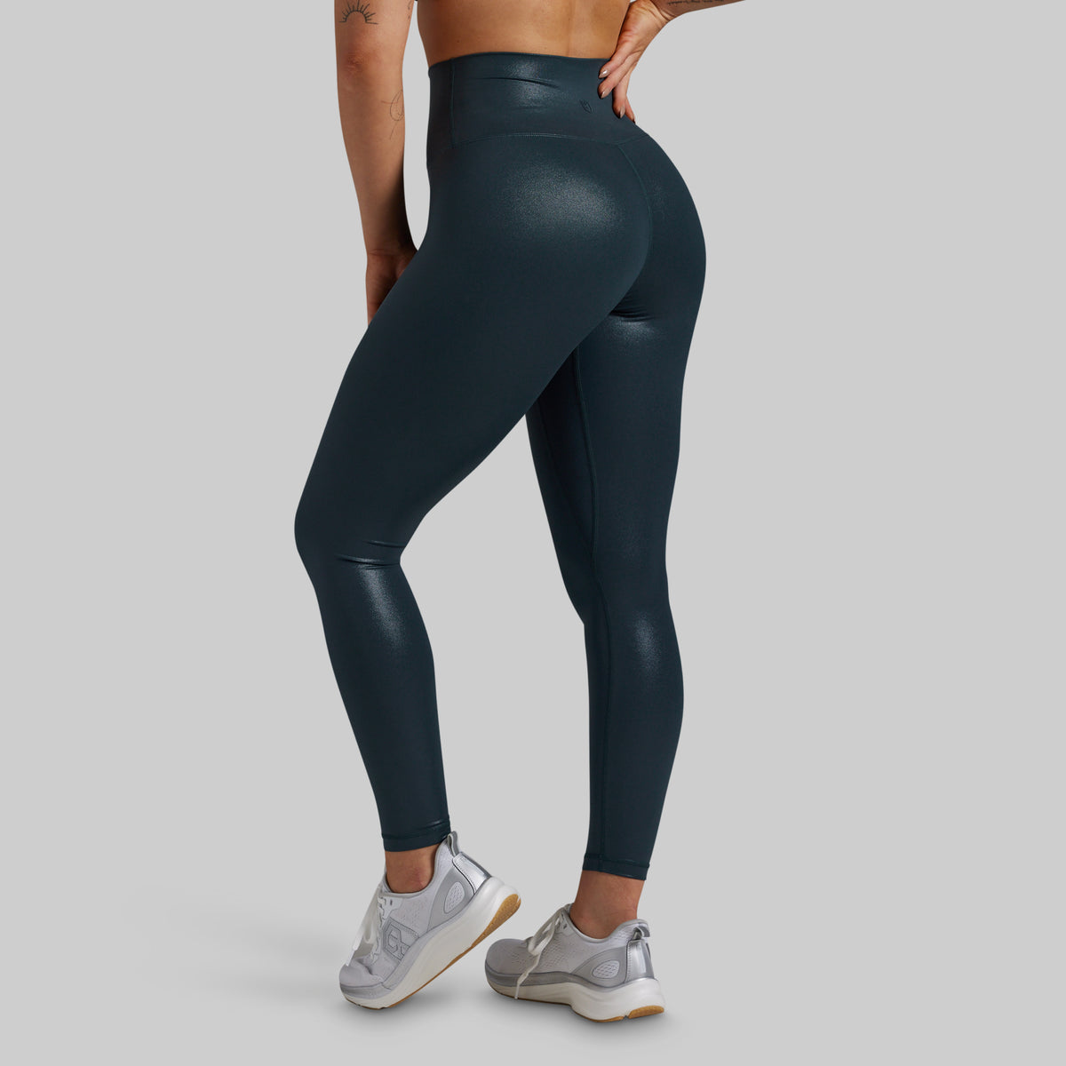 A person wearing high-waisted, shiny dark teal leggings, shown from the back and side against a light gray studio background. The model is also wearing light gray sneakers.