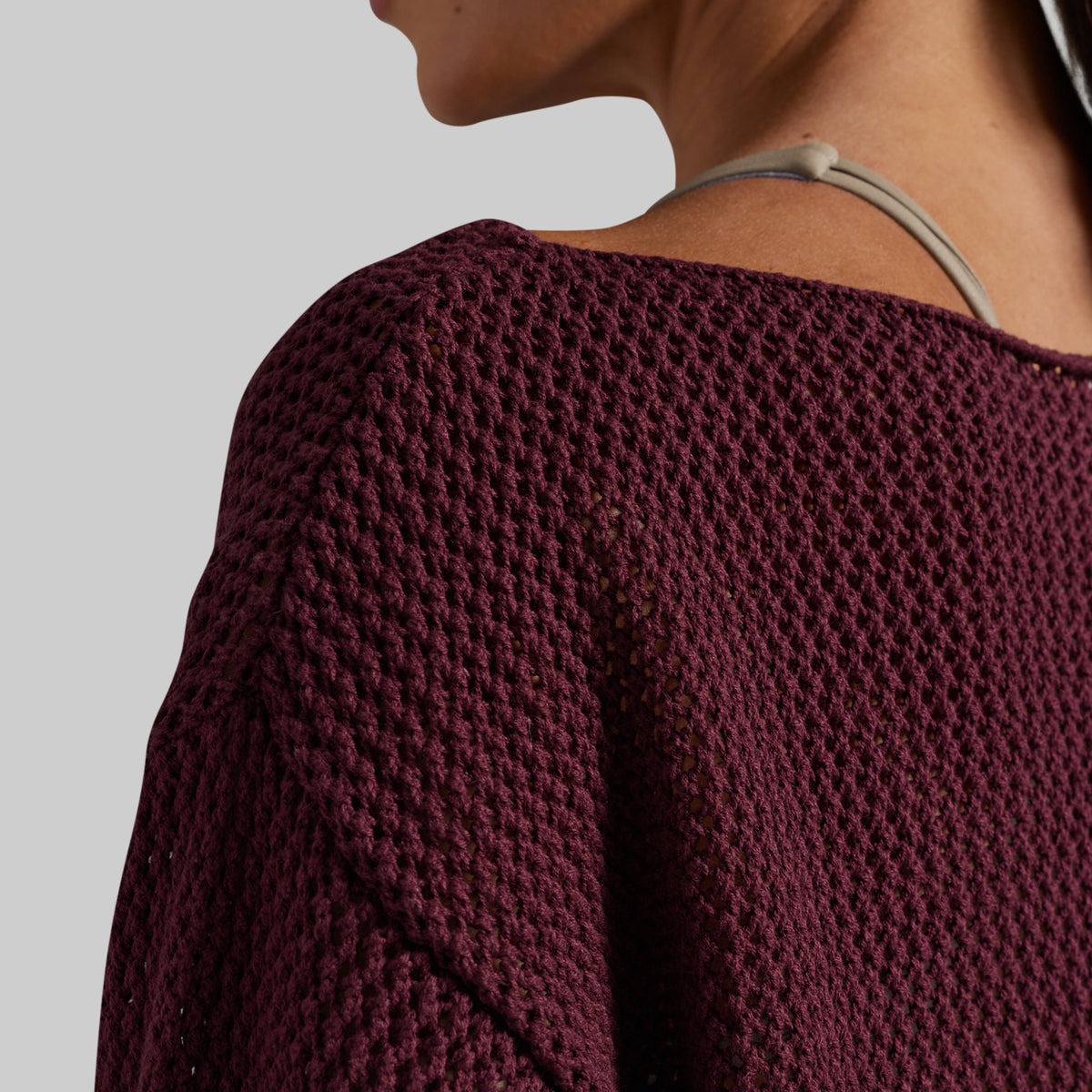 Close-up of a person wearing a chunky knit burgundy sweater, showing the textured fabric and shoulder strap detail against a light gray background.
