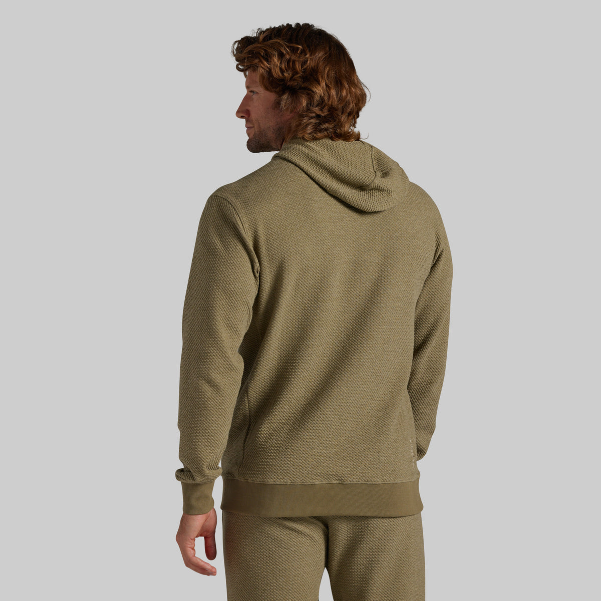 A man shown from the back wearing a textured olive green hooded sweatshirt and matching sweatpants against a solid light gray background.