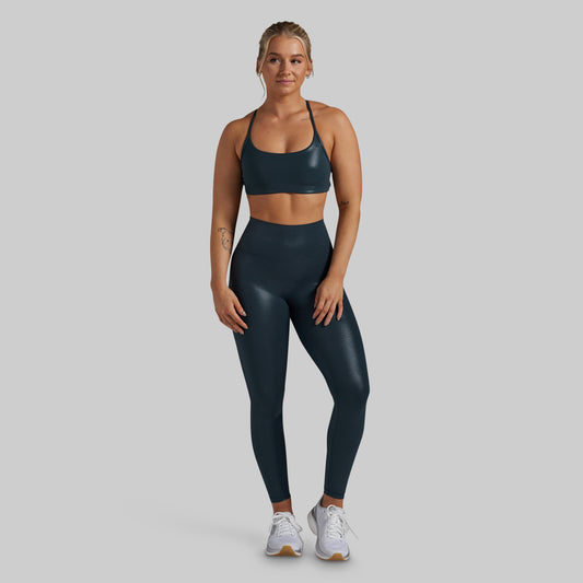 A woman modeling a matching dark teal athletic set consisting of a sports bra and high-waisted leggings, paired with white sneakers, against a solid light gray background.