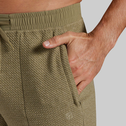 Close-up of a person wearing textured olive green athletic joggers, with one hand tucked into the side pocket. A small tonal geometric logo is visible near the lower leg on a light gray background.