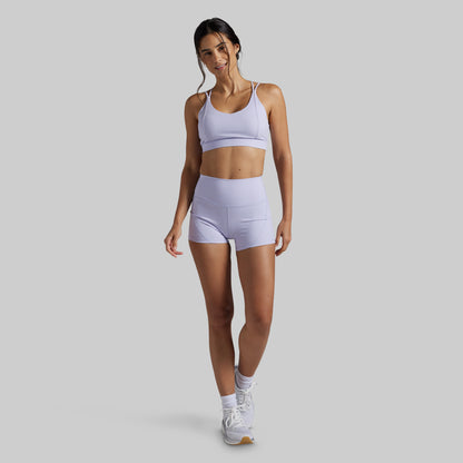 A woman modeling a matching light lavender athletic set consisting of a sports bra and high-waisted shorts, paired with white sneakers, against a solid light gray background.