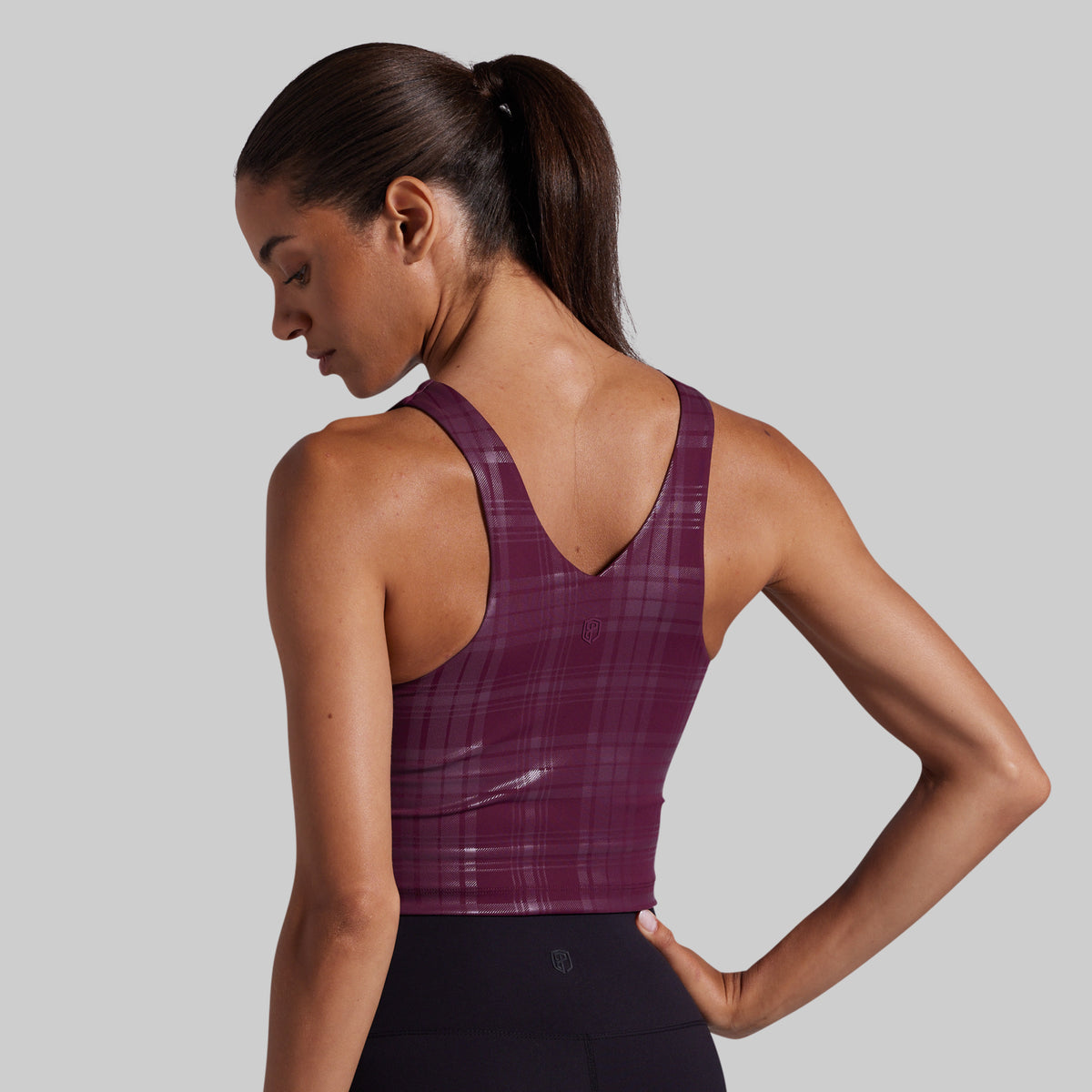 A woman shown from the back wearing a plum purple athletic sports bra with a subtle plaid pattern, paired with black leggings, against a solid gray studio background.