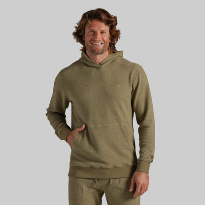 A male model wearing textured olive green hooded sweatshirt and matching sweatpants, standing against a solid gray studio background.