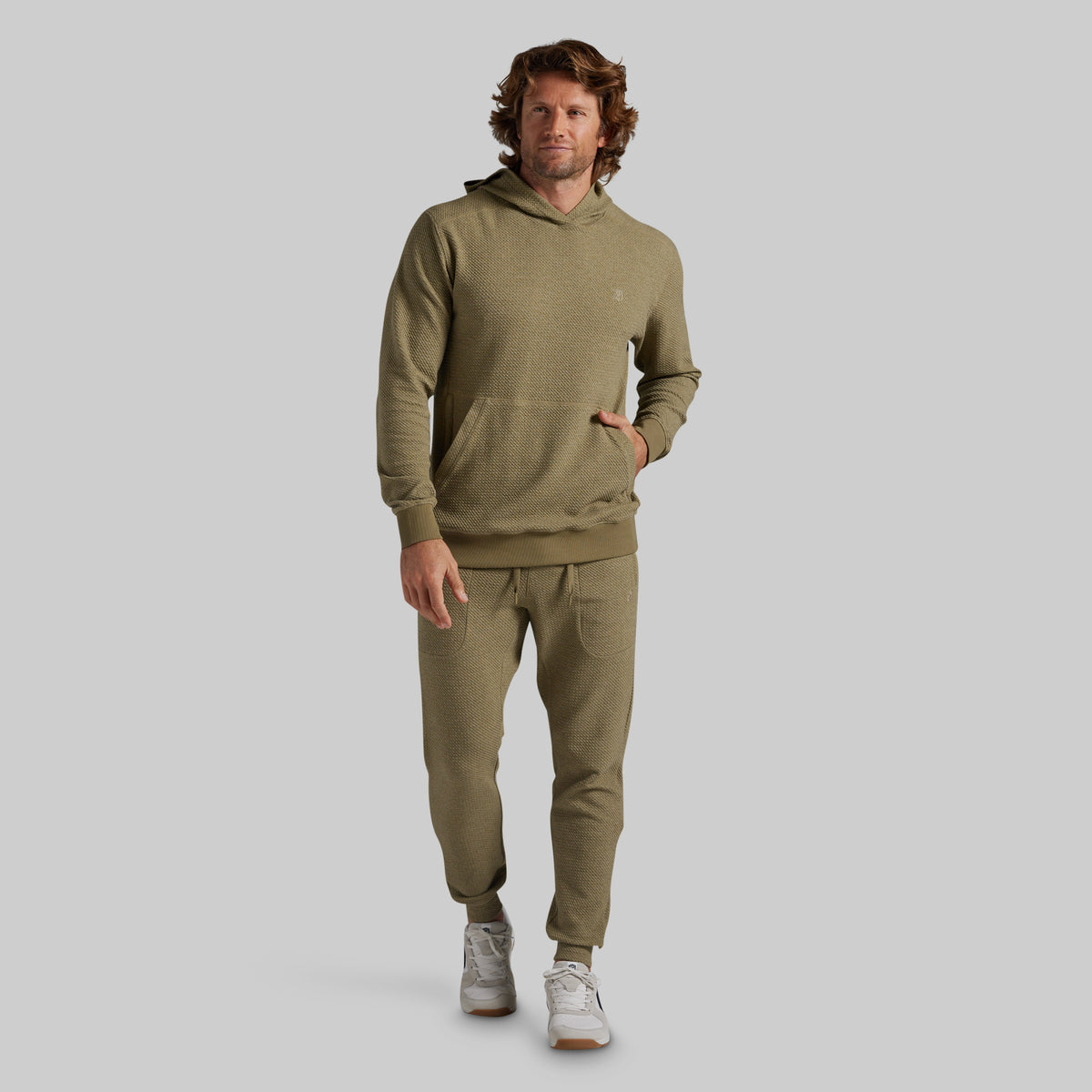 A man modeling a matching olive green hoodie and jogger sweatpants set against a solid gray studio background.