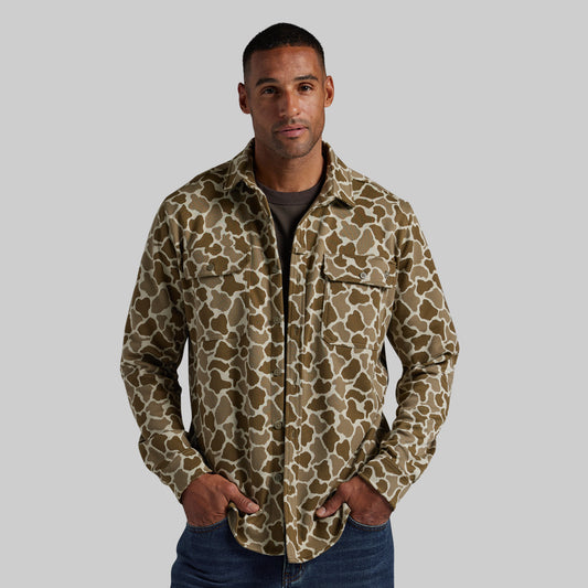 A man wearing a long-sleeve button-up shirt with a brown and beige animal print pattern, paired with a dark gray t-shirt and blue jeans, against a solid gray background.