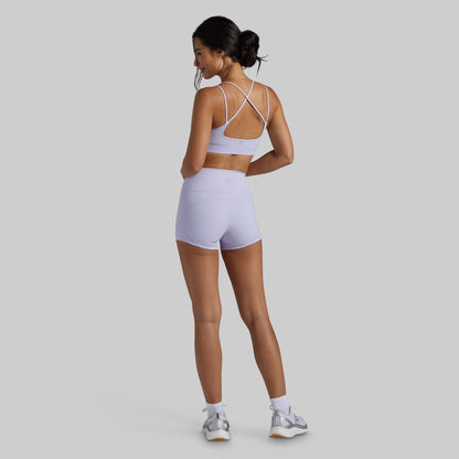 A woman wearing a light lavender athletic set consisting of a sports bra and matching high-waisted shorts, shown from the back against a solid gray background.