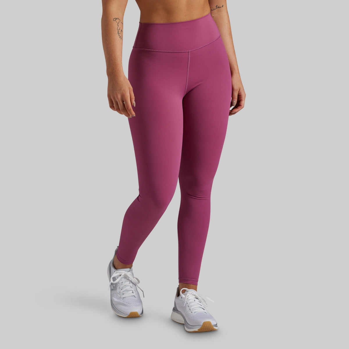 A model wearing high-waisted, form-fitting leggings in a dusty rose or mauve shade, paired with white athletic sneakers, shown from the waist down against a solid light gray background.