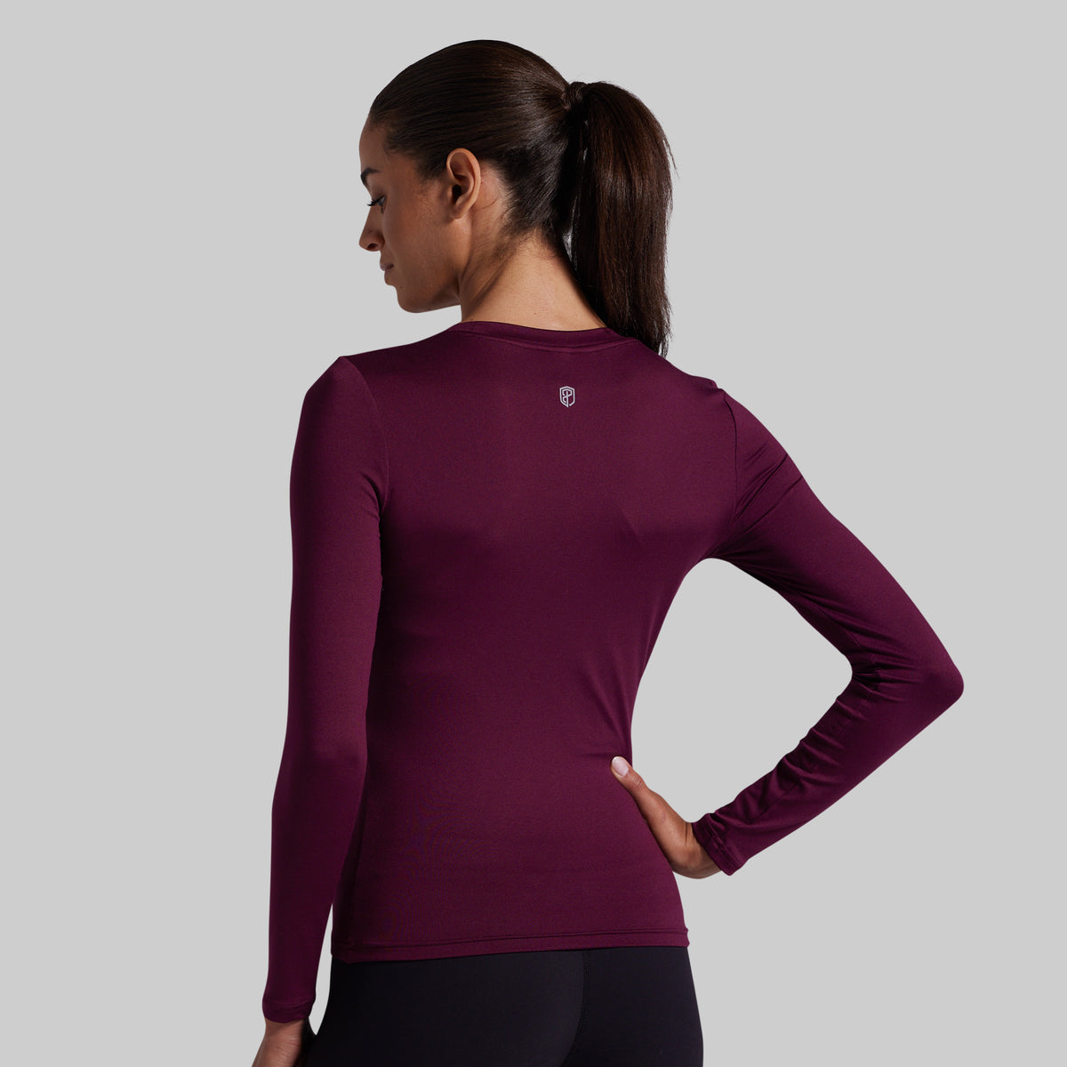 A woman shown from the back wearing a long-sleeve athletic top in deep burgundy, paired with black leggings, against a solid gray studio background.