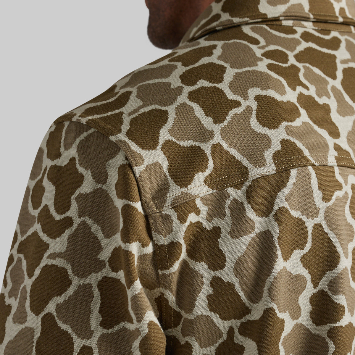 Close-up of a person wearing a collared shirt with a brown and beige animal print pattern, showing the shoulder and upper back against a light gray background.