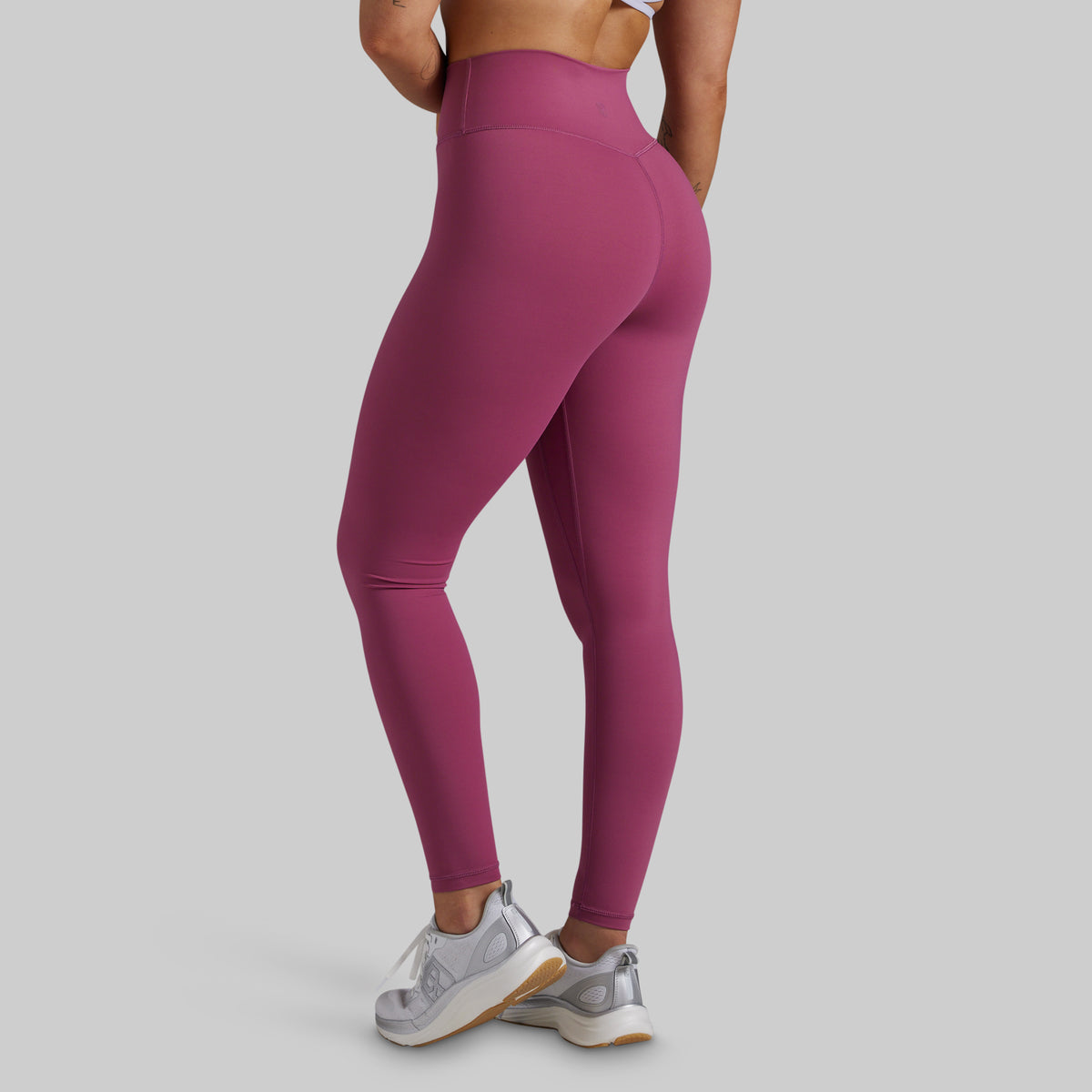 A model from the back wearing high-waisted, form-fitting leggings in a dusty magenta pink color, paired with white and silver sneakers, against a solid light gray background.