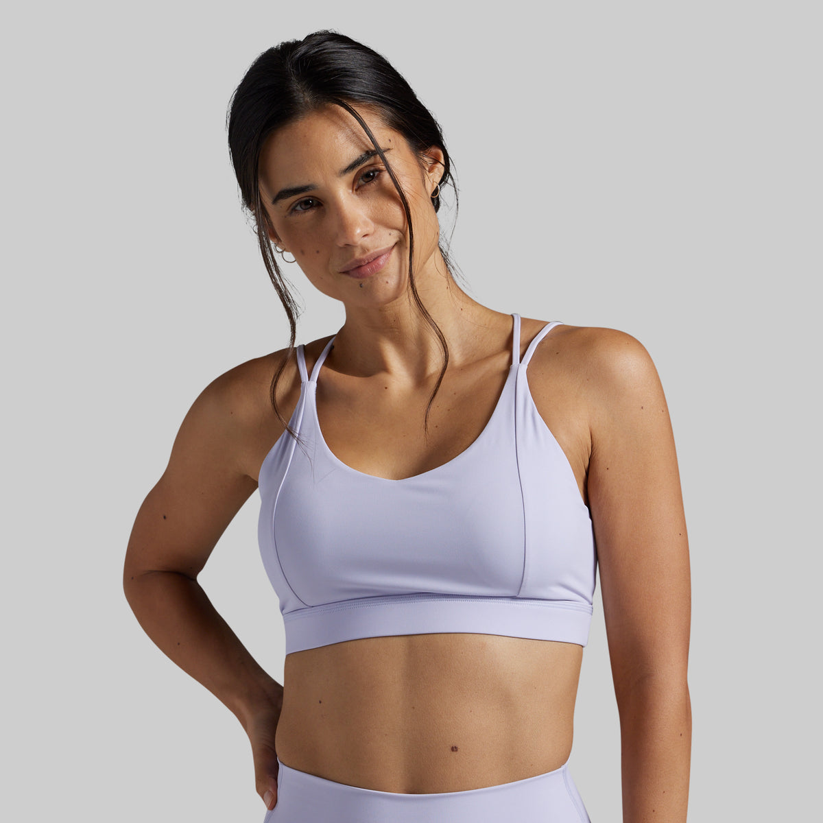 A woman modeling a light lavender sports bra against a solid gray studio background.