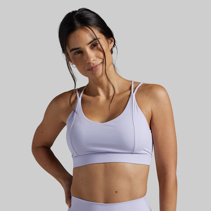 A woman modeling a light lavender sports bra against a solid gray studio background.
