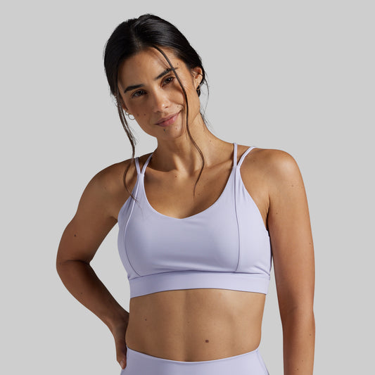 A woman modeling a light lavender sports bra against a solid gray studio background.