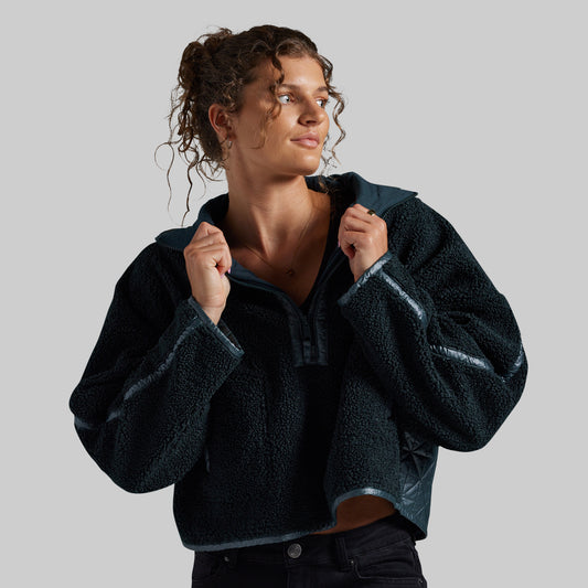 A woman wearing a dark teal fleece pullover jacket with contrasting shiny dark teal trim, posing against a solid gray studio background.