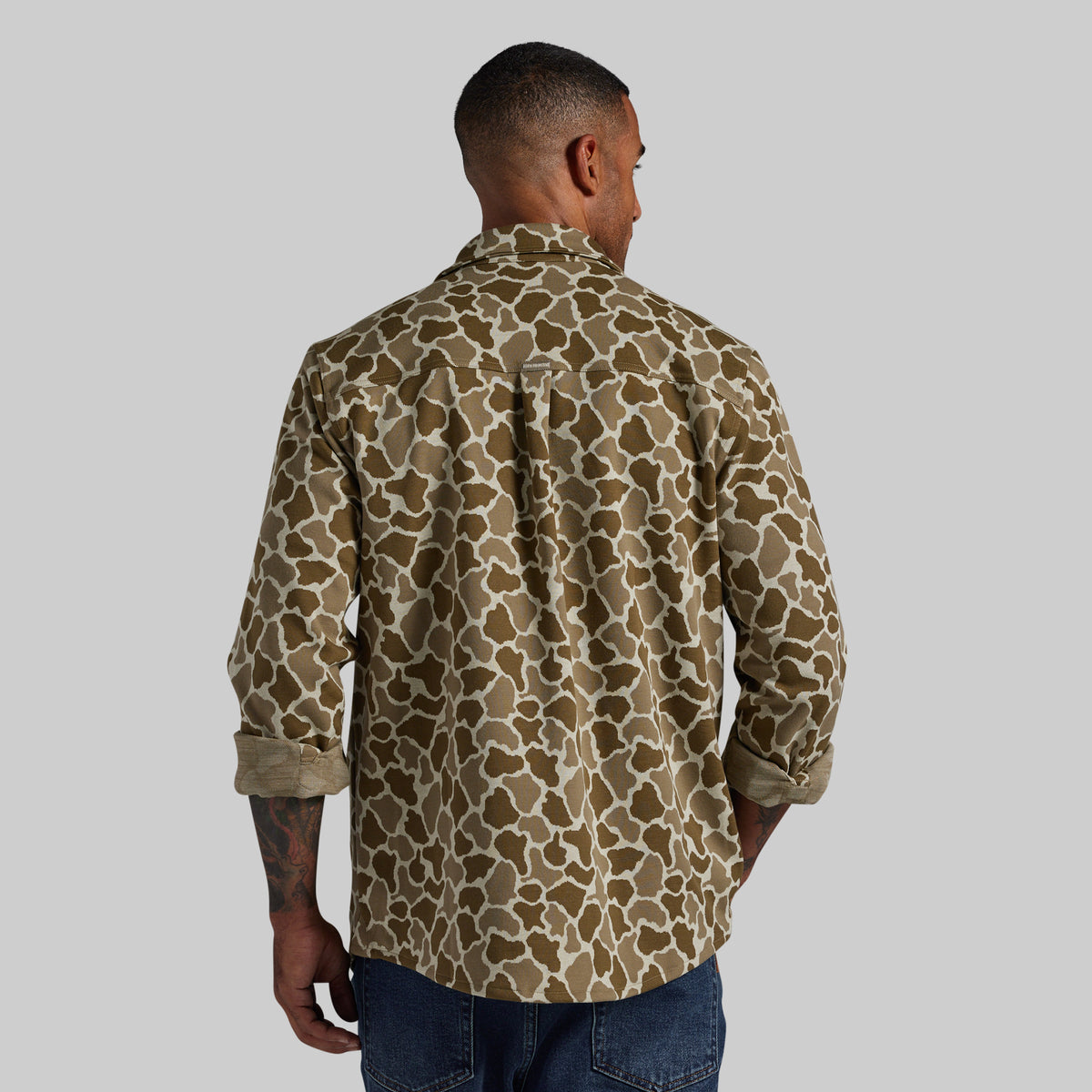 A man viewed from behind wearing a long-sleeve button-up shirt with a brown and beige organic animal-style pattern, paired with blue jeans, against a solid gray background.