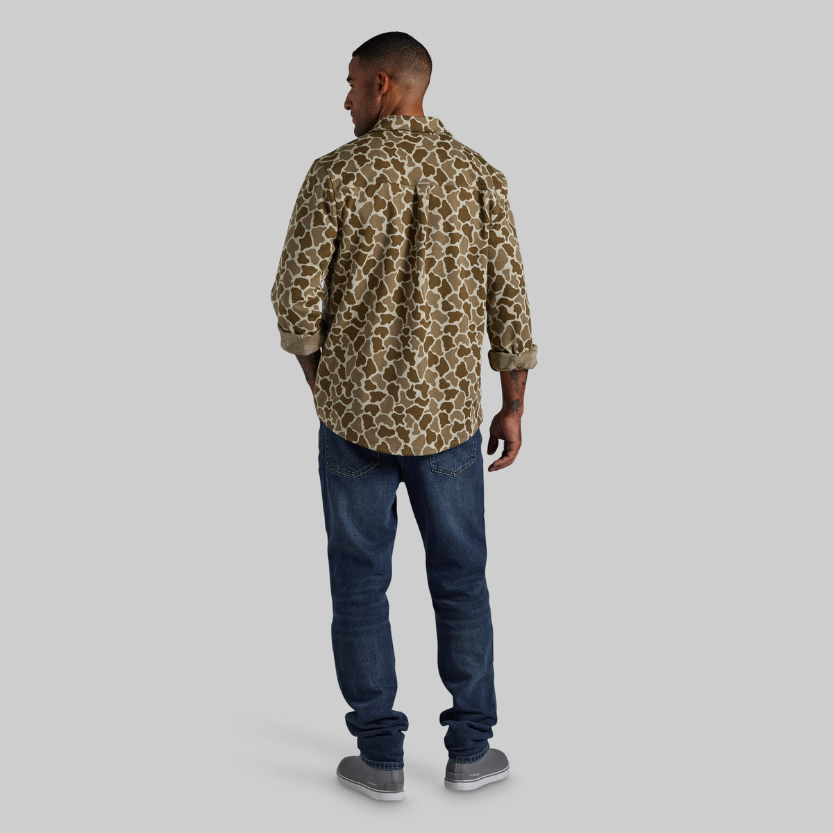 A man viewed from behind wearing a long-sleeve button-up shirt with a brown and beige animal print pattern, paired with dark blue jeans and gray sneakers, against a solid light gray background.