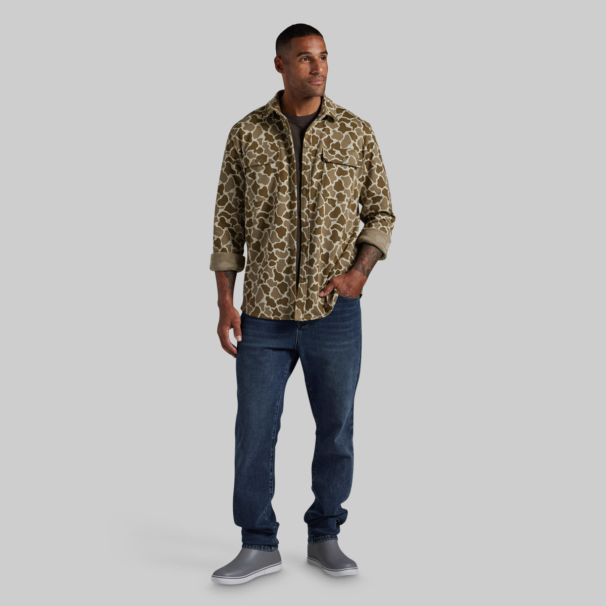 A man modeling a flannel with a brown and beige frog camo print pattern, paired with dark blue jeans and gray slip-on shoes, against a solid gray background.