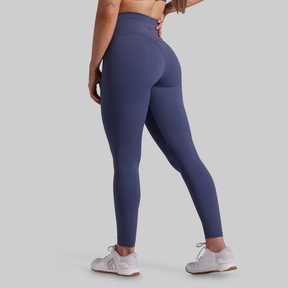 A woman wearing high-waisted, full-length athletic leggings in a muted slate blue color, shown from a rear three-quarter angle against a solid light gray background.