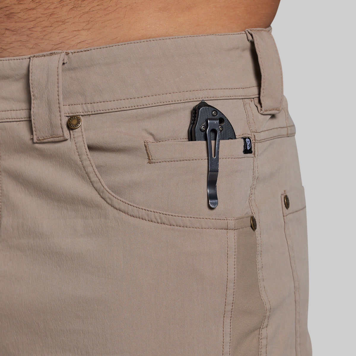 Close-up of khaki pants worn by a person, showing the back pocket with a black clip holding a small black accessory, against a light gray background.