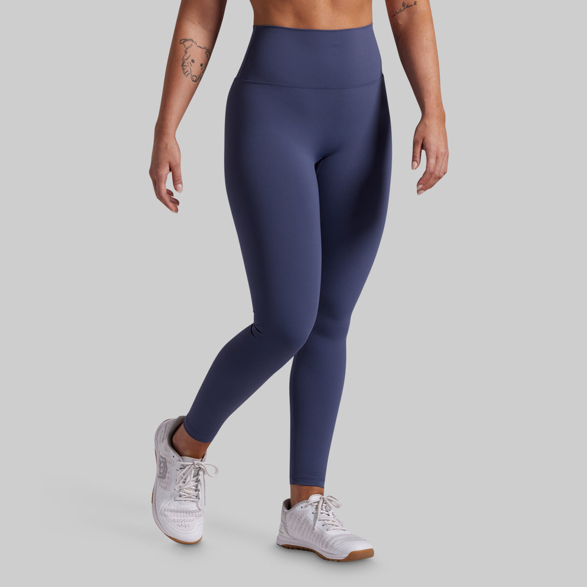 A model wearing high-waisted, full-length athletic leggings in a solid blue color, paired with white sneakers, shown from the waist down against a solid light gray background.