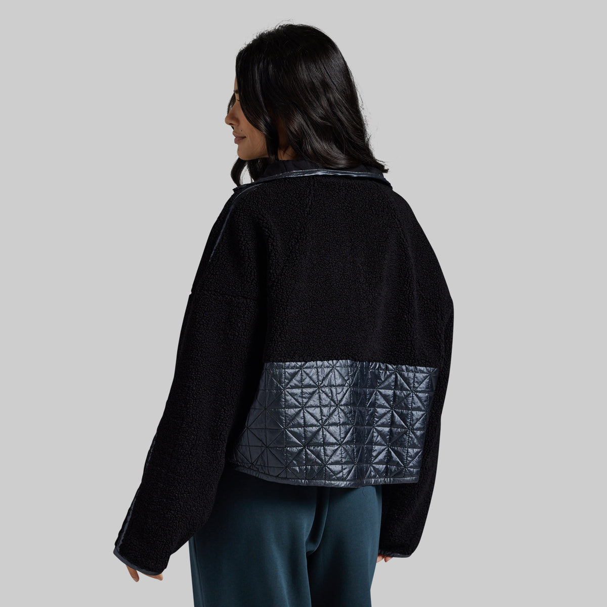 A woman shown from the back wearing a black cropped jacket with a dark gray quilted geometric patterned panel on the lower back, paired with dark teal pants, against a solid light gray background.
