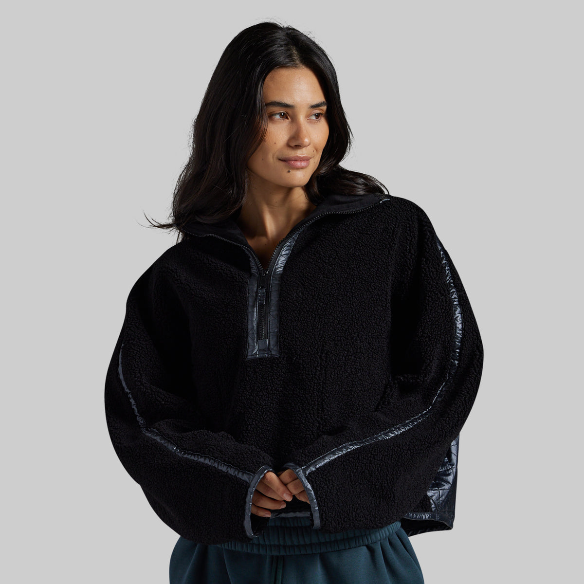 A woman wearing a black fleece half-zip pullover with shiny dark gray contrast trim on the collar, zipper, and cuffs, photographed against a solid gray studio background.