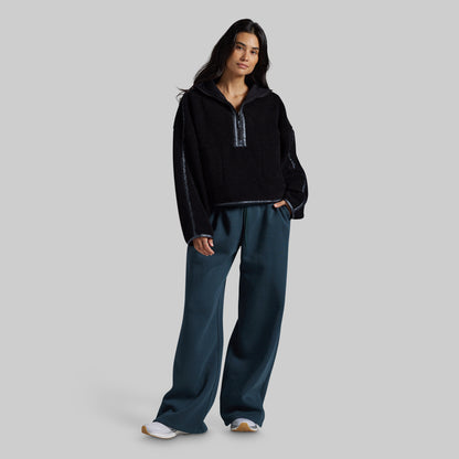 A woman modeling a black fleece half-zip pullover with textured knit sleeves and dark teal wide-leg sweatpants, standing against a solid gray studio background.