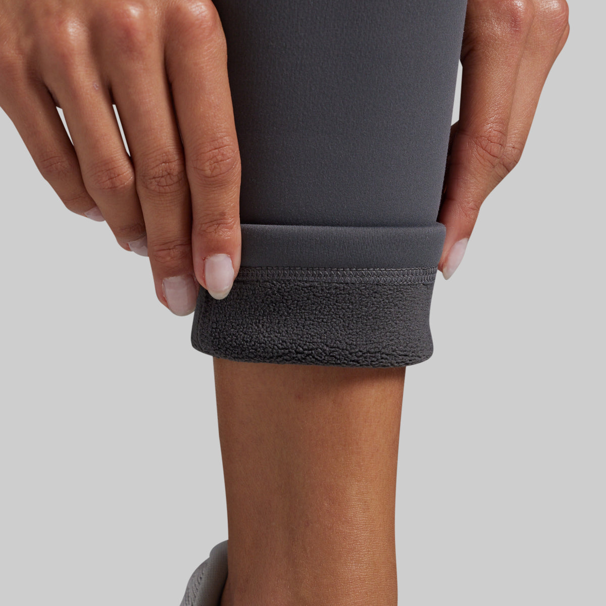 Close-up of a person's hands and lower leg pulling up the cuff of dark gray leggings to reveal a soft, fleece-lined interior.