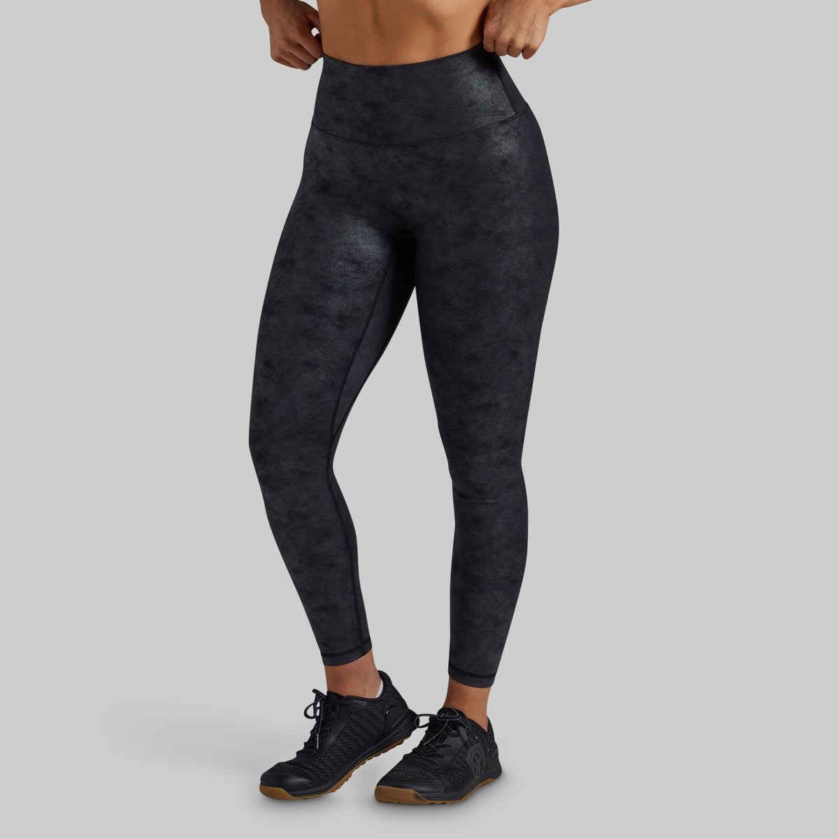 A model wearing high-waisted, full-length athletic leggings with a dark charcoal gray and black mottled pattern, paired with black athletic shoes, against a solid light gray background.