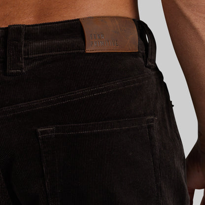 Close-up of the back waistband of dark brown corduroy pants worn by a model, showing a brown leather brand patch that reads 'BORN PRIMITIVE'.