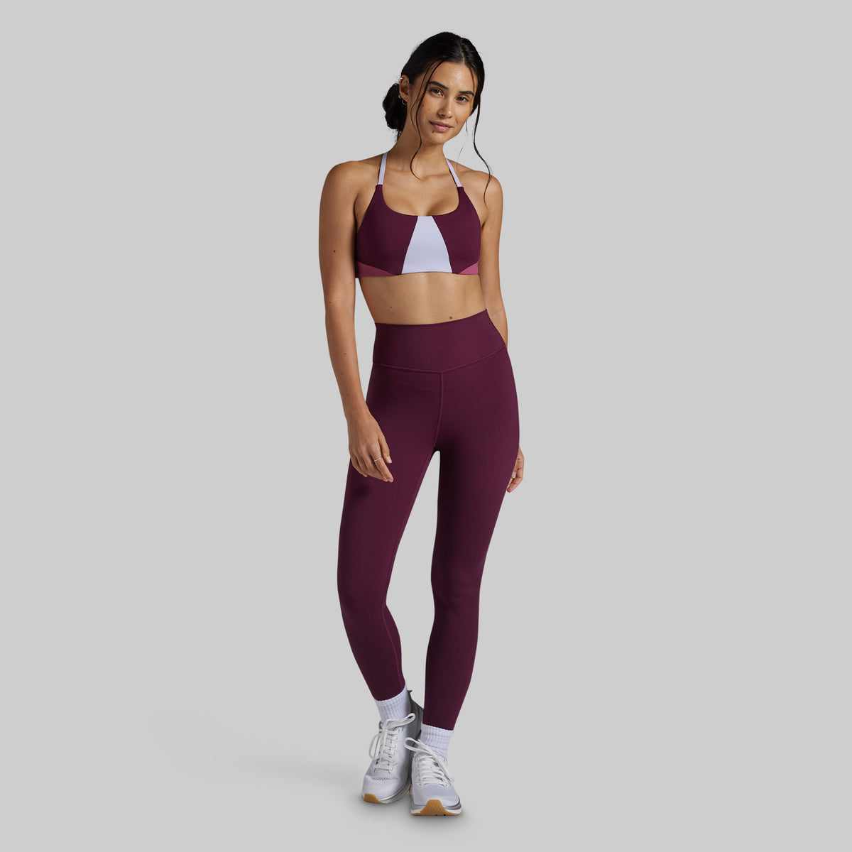 A woman modeling a matching athletic set consisting of a maroon sports bra with a white front panel and high-waisted maroon leggings, paired with white sneakers, against a solid gray background.