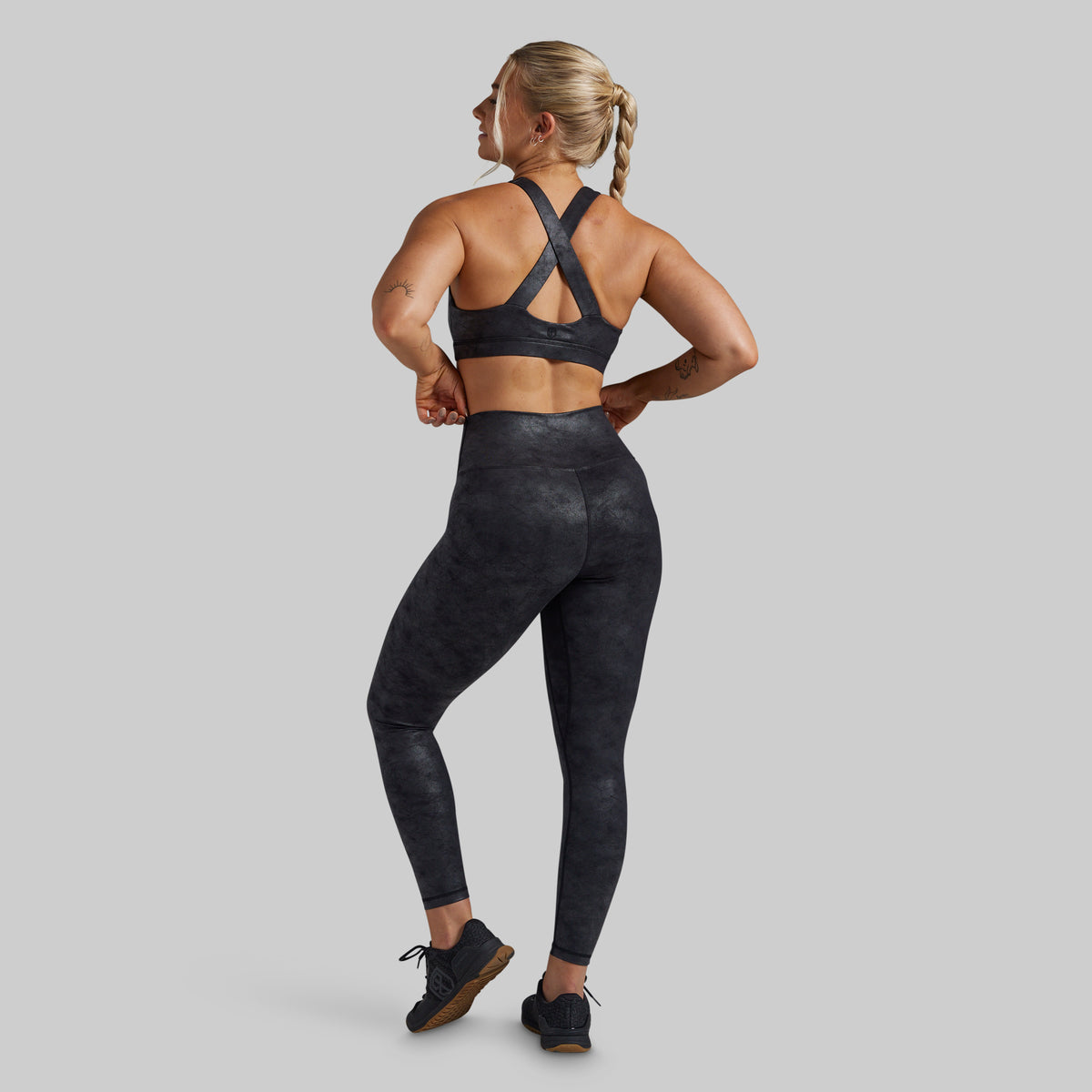 A woman shown from the back wearing a matching black athletic set, featuring a sports bra with crisscross straps and high-waisted leggings with a subtle mottled pattern, set against a solid gray background.