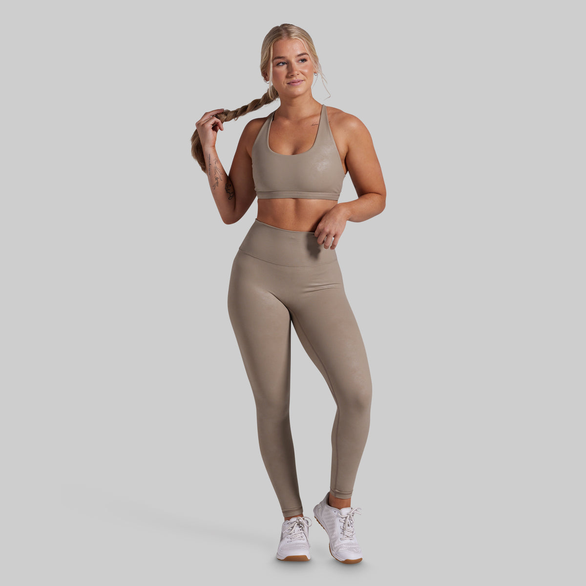 A woman modeling a matching athletic set consisting of a sports bra and high-waisted leggings in a taupe color, paired with white sneakers, against a solid light gray background.
