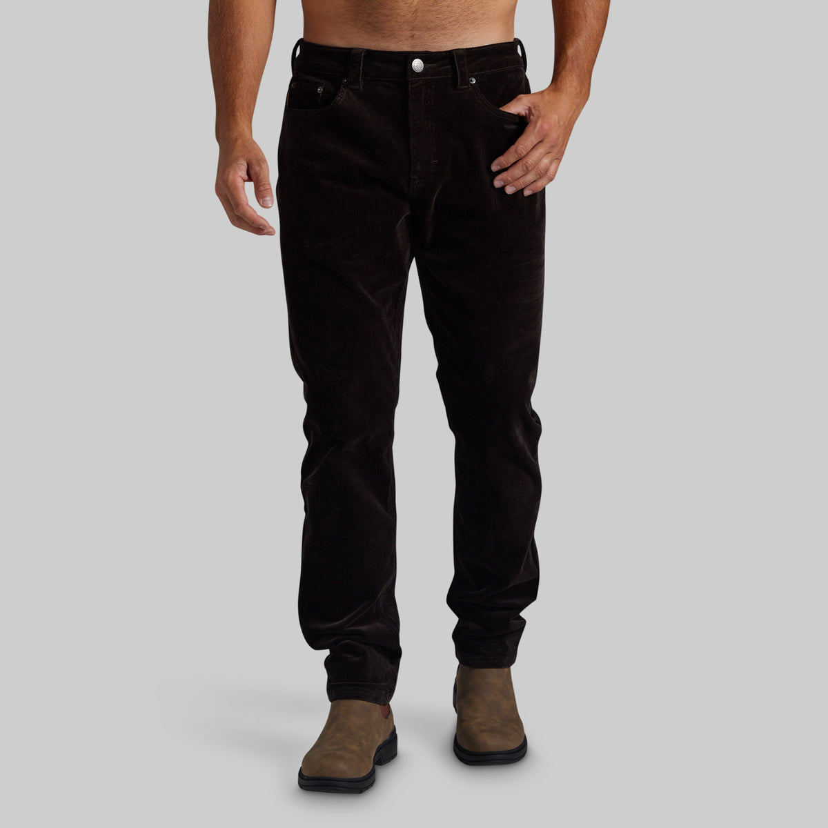 A person modeling slim-fit dark brown corduroy pants with brown suede boots, shown from the waist down against a plain light gray studio background.