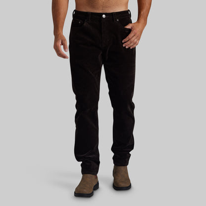 A person modeling slim-fit dark brown corduroy pants with brown suede boots, shown from the waist down against a plain light gray studio background.