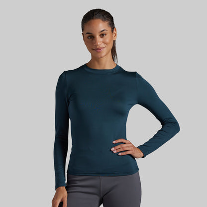 A woman modeling a dark teal long-sleeve athletic top, paired with gray leggings, against a solid gray background.