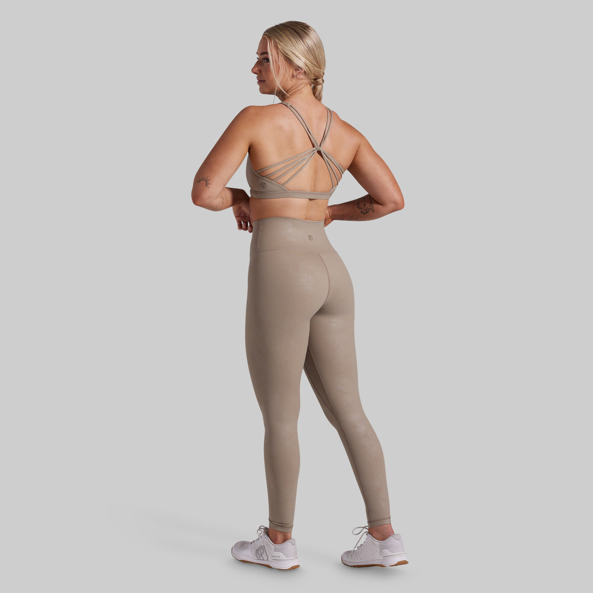 A woman is shown from the back wearing a matching beige sports bra and high-waisted leggings set, paired with white sneakers, against a solid light gray background.