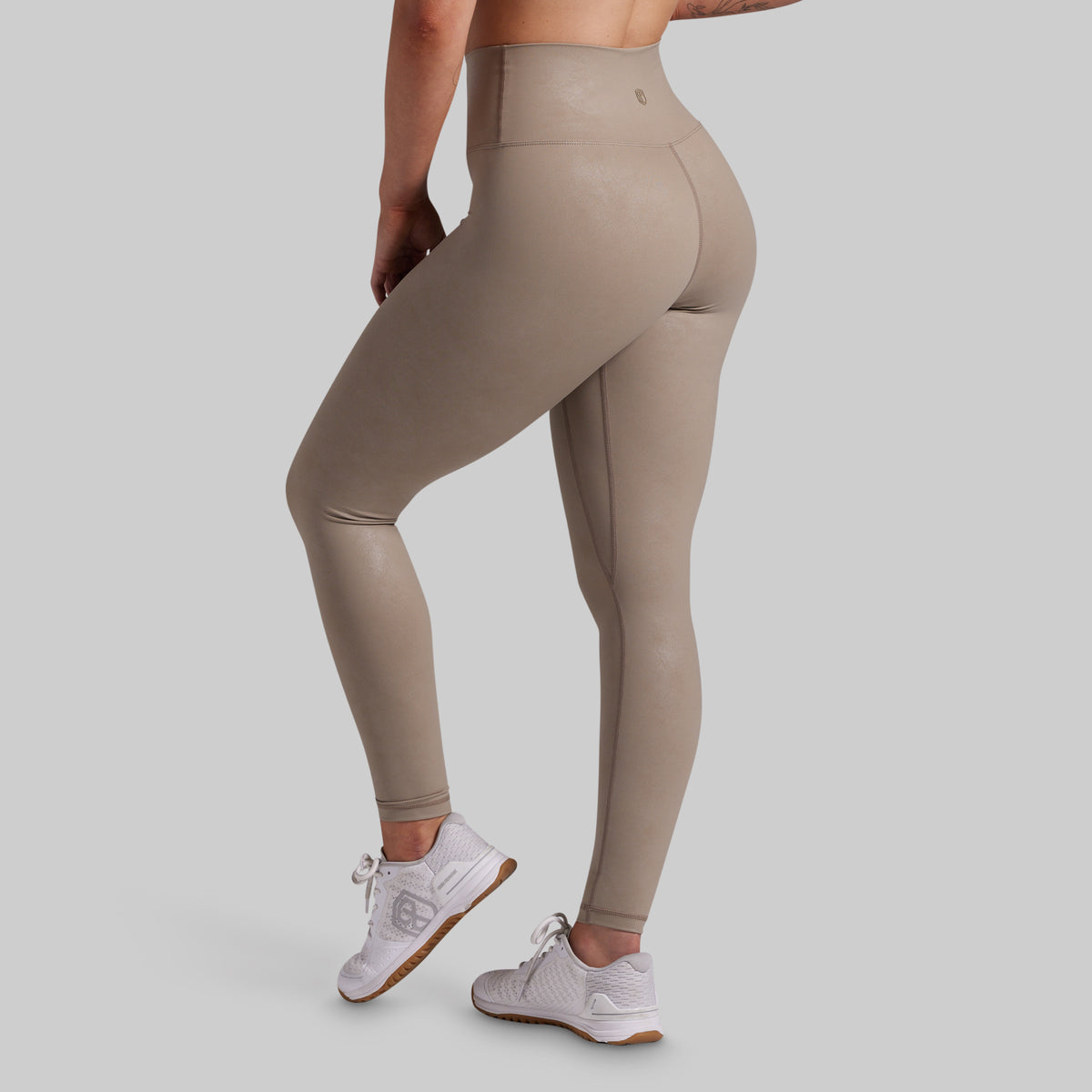 A person wearing high-waisted, shiny taupe-beige leggings and white sneakers, shown from the side and back against a light gray studio background.
