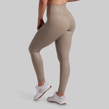 A person wearing high-waisted, shiny taupe-beige leggings and white sneakers, shown from the side and back against a light gray studio background.