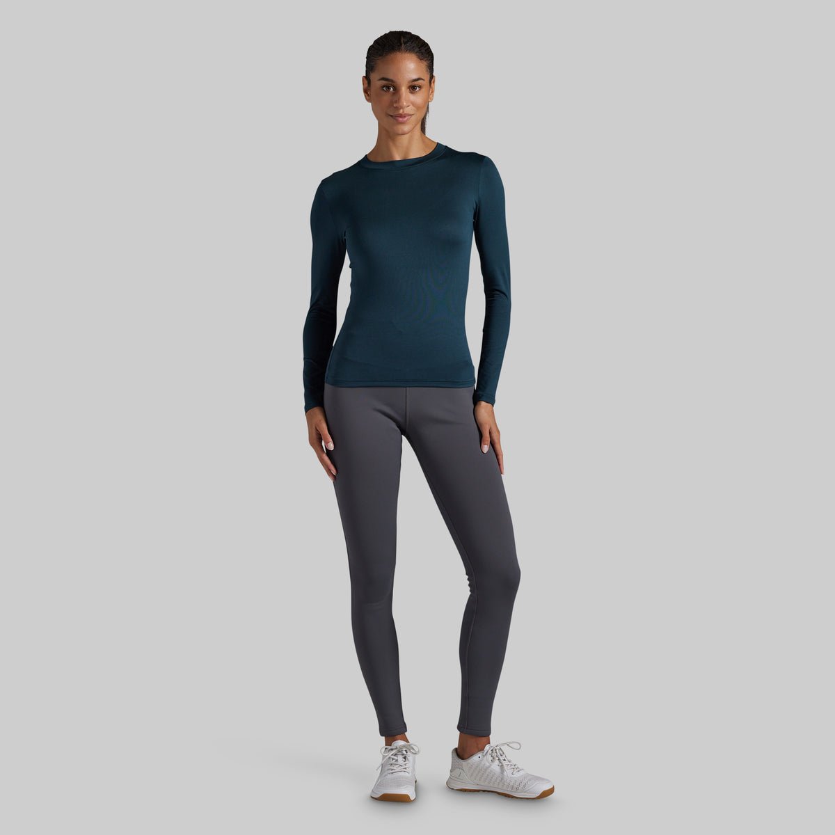 A woman modeling a dark teal long-sleeve athletic top and dark gray leggings with white sneakers, standing against a solid light gray background.