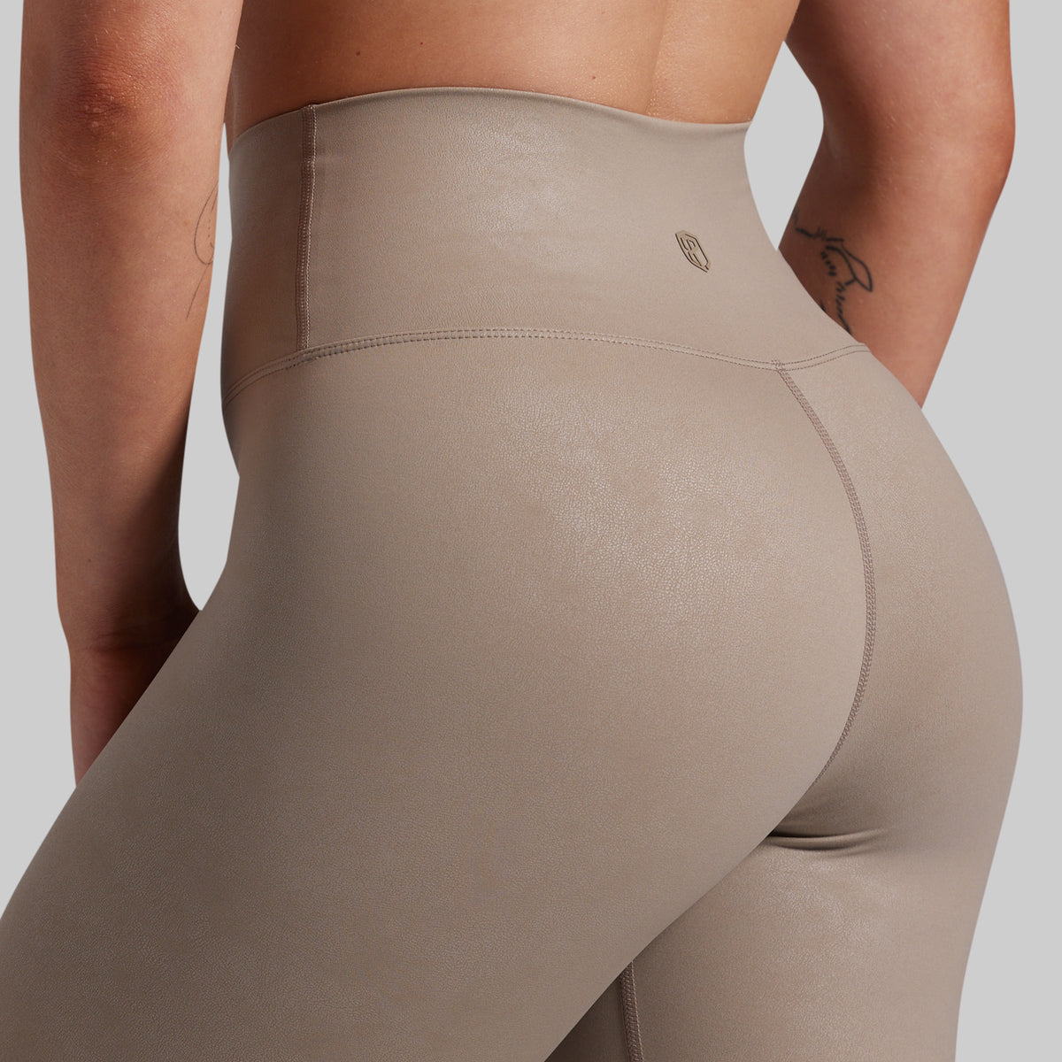Close-up rear view of a person wearing high-waisted taupe leggings against a light gray studio background.
