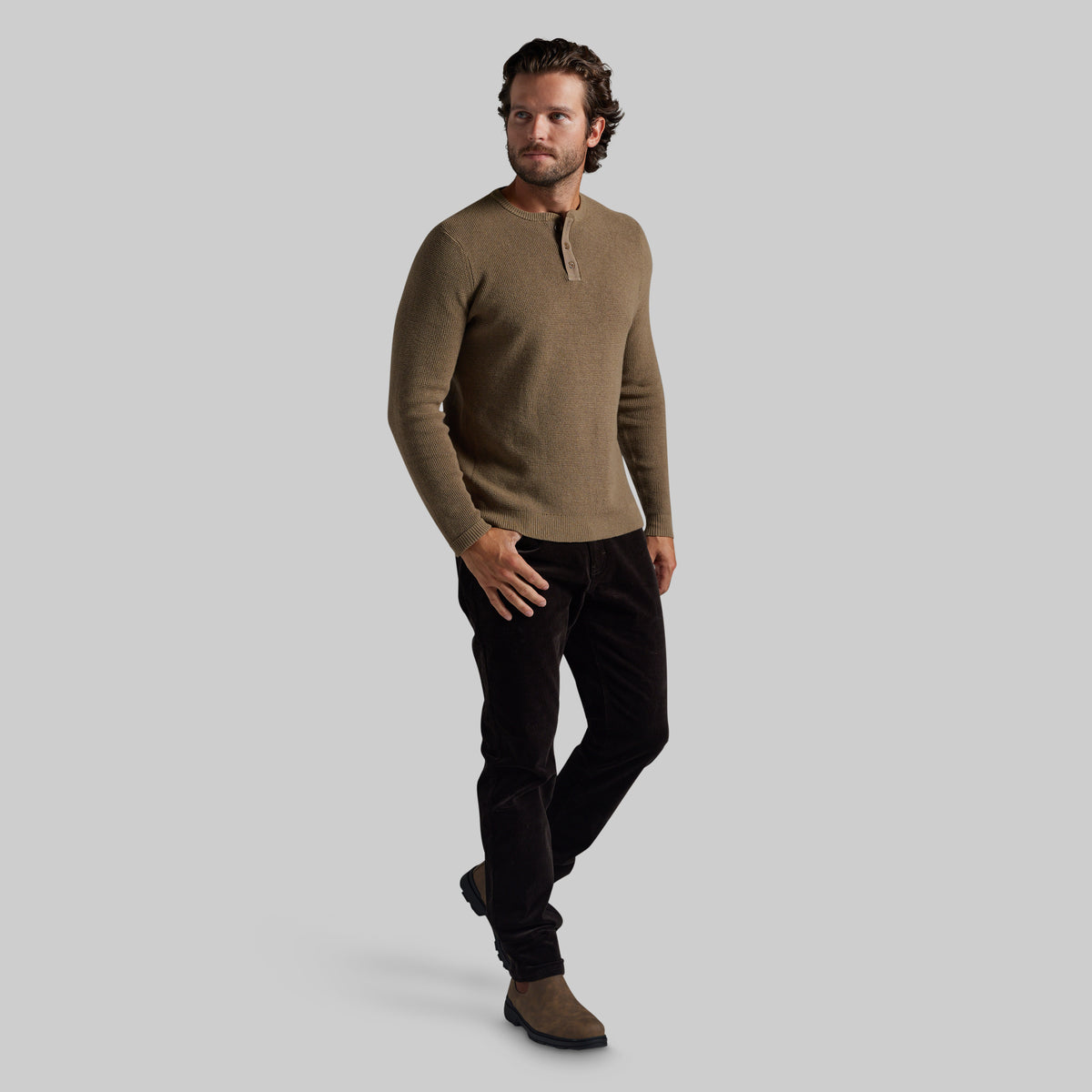 A man modeling a long-sleeve henley sweater in a warm taupe-brown color, paired with black pants and brown boots, against a solid light gray background.